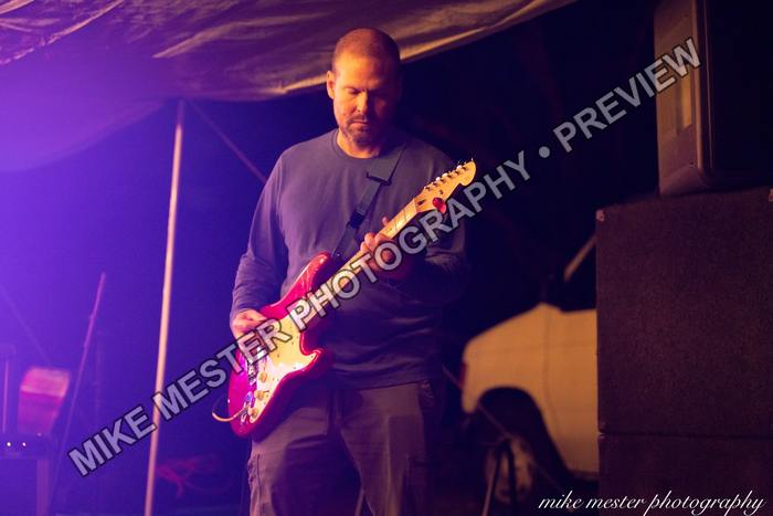 Guitarist playing a red electric guitar beneath purple stage light.